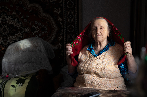 Grandmother sits at the table and puts a headscarf on her head.