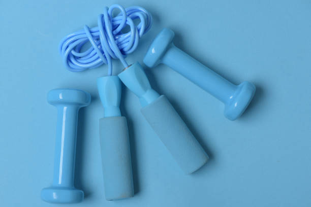 Jump rope and barbells placed next to each other. Fit shape and sport concept. Health regime and fitness symbols. Dumbbells and skipping rope in cyan color on blue background, top view