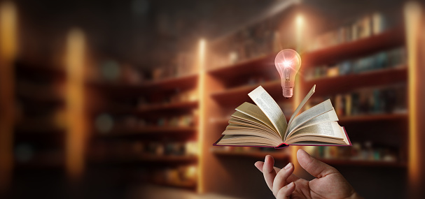 The hand shows a book and a burning light bulb above it against the background of shelves with books.