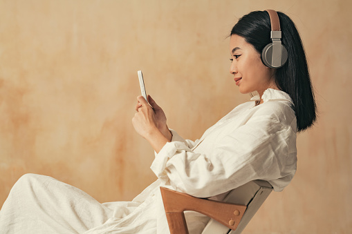 Side view closeup of korean girl in headphones looking at smartphone screen, watching podcast or reading e-book, listening to music with alpha rhythms in her wireless headphones on brown background