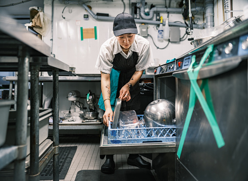 Young Korean female chef working in restaurant kitchen. She is wearing usual kitchen attire with apron and baseball cap. Interior of industrial kitchen of a busy restaurant.