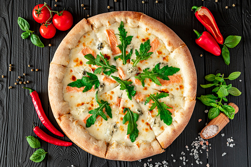 Delicious hot pizza with salmon, arugula and different spices on wooden table ready to eat. Tasty and big pizza with different types of meat. Top view of big pizza