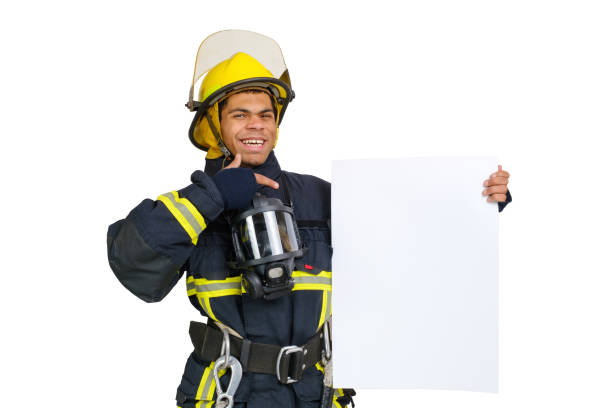 Young smiling African American fireman wearing uniform and helmet looking to the camera and holding blank paper sheet in hand and finger pointing. Copy space for text, isolated on white background