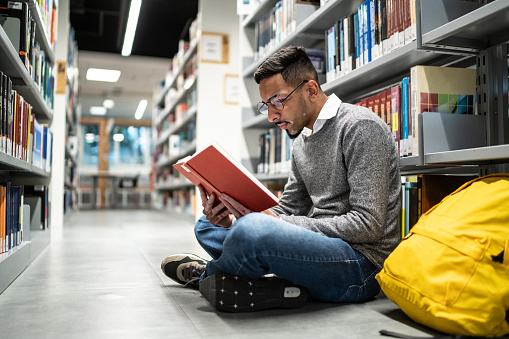 teenage student sits on floor of library or bookstore and reads book with enthusiasm