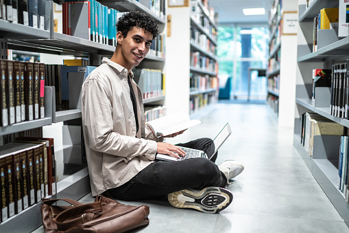 Portrait of a young student man using a laptop sitting on ground at university library