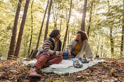 Smiling Hispanic couple in mid 40s wearing warm clothes, sitting on a blanket and having tea time in the woods.