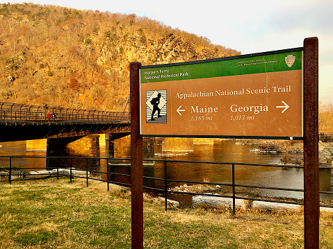 Harpers Ferry, West Virginia, USA - December 2, 2017: A sign shows which direction to take the Appalachian National Scenic Trail as two people walk the trail across a railroad bridge crossing the Potomac River from the Maryland side of the river.