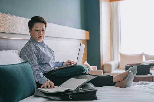 Asian Chinese businesswoman working in hotel room typing using laptop reading documents on bed