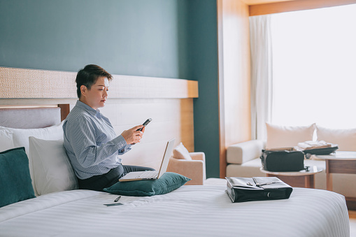 Asian Chinese businesswoman working in hotel room text messaging with smart phone on bed