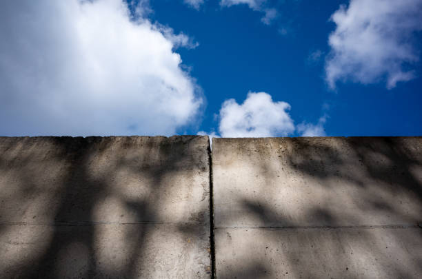 A massively high concrete security boundary wall topped with barbed wire on a blue sky background - 3D render