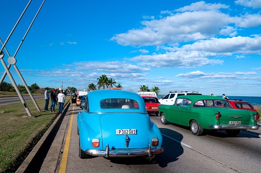 Havana, Cuba – February 01, 2022: a long line of cars stopped next to the sea in a road under a sunny day, the foreground with American classic cars, people waiting at the road side