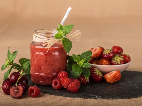 A closeup shot of a juicy red smoothie near a bunch of cherries, raspberries, and strawberries