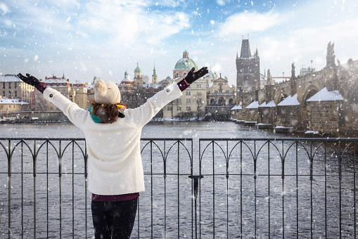 A happy tourist woman in winter clothing enjoys the view of the skyline of Prague with Charles Bridge and old town with snow