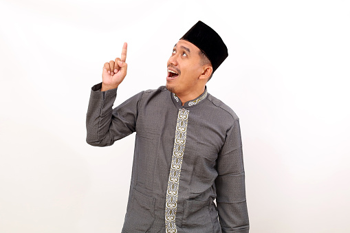 Surprised asian muslim man standing while pointing something above him. Isolated on white background