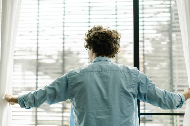 istock Young man from behind opens the window curtains and looks out from his workplace 1453442632