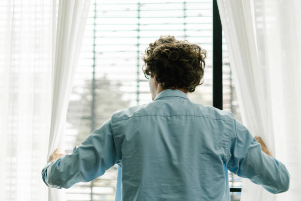 istock Young man from behind, who opens the window curtains and looks out from his workplace 1453443301