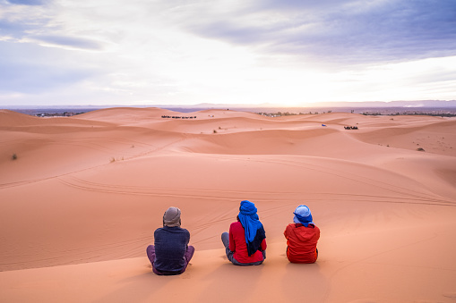 Three adventure traveler friends watch the sunset from a dune in the Sahara desert. A trip with friends strengthens the friendship and improves the level of trust and closeness.