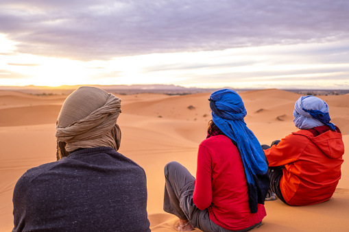 Three adventure traveler friends watch the sunset from a dune in the Sahara desert. A trip with friends strengthens the friendship and improves the level of trust and closeness.