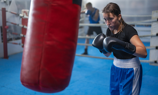 Teenage girl exercising at boxing with black boxing gloves on punching bag with another kids from boxclub. Combat sport and healthy lifestyle concept.