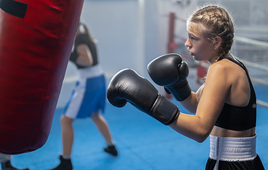 Teenage girl exercising at boxing with black boxing gloves on punching bag with another kids from boxclub. Combat sport and healthy lifestyle concept.