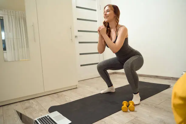 Smiling cheerful athletic female performing squats on exercise mat before laptop in bedroom