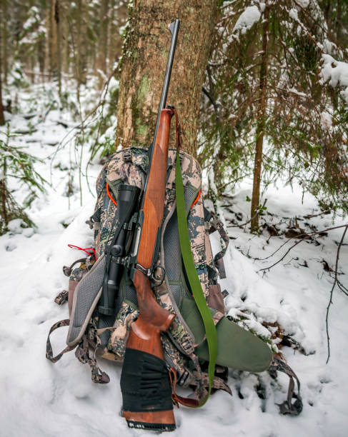 Hunting carbine stands near tree trunk next to camouflage backpack on winter hunt. Rifle with an optical sight lies on hunting backpack in snowy forest.