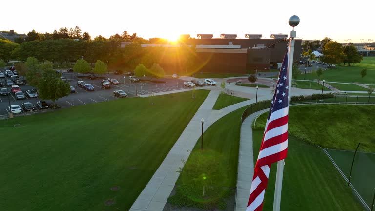 Glare of the sun over a high school building. Aerial shot of American flag in the foreground. Well-maintained academic campus.