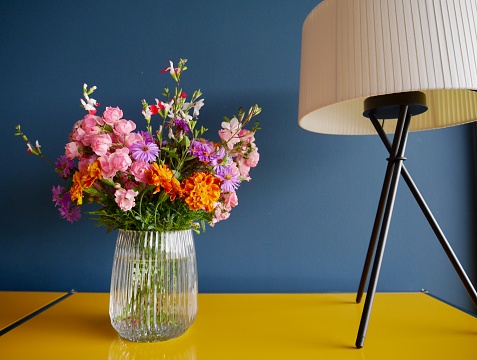 olorful garden flowers in glass vase on yellow sideboard against petrol blue wall with stylish table lamp.