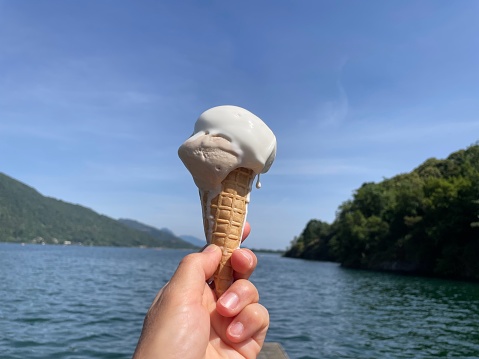 Female hand holding ice-cream cone in hand, beautiful Mergozzo lake in the background. Piedmont, Italy.
