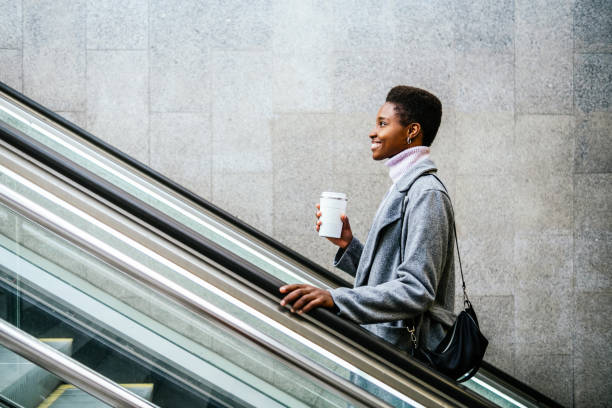 Cheerful black woman with coffee riding escalator Side view of stylish African American female in stylish coat and sweater with cup of coffee and bag smiling and riding moving stairs in city escalator stock pictures, royalty-free photos & images