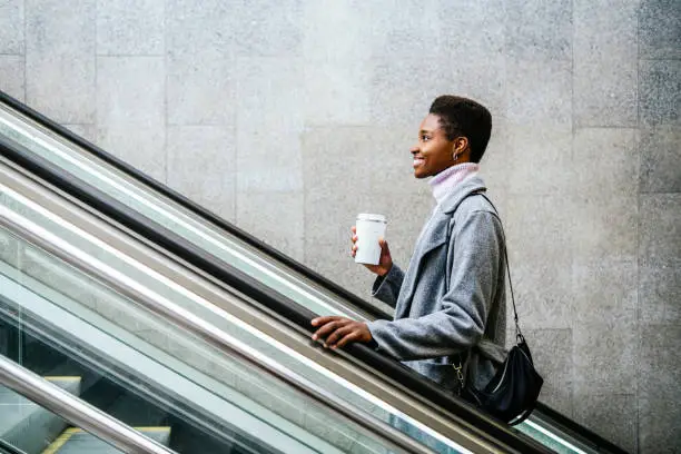 Side view of stylish African American female in stylish coat and sweater with cup of coffee and bag smiling and riding moving stairs in city