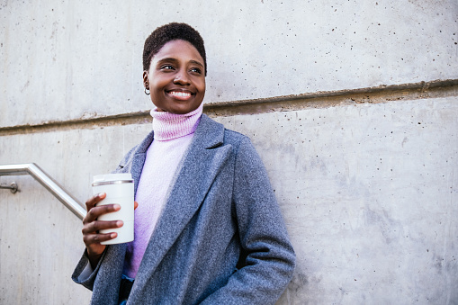 Happy African American female in stylish coat and sweater with zero waste cup of coffee smiling and looking away while leaning on concrete wall on city street