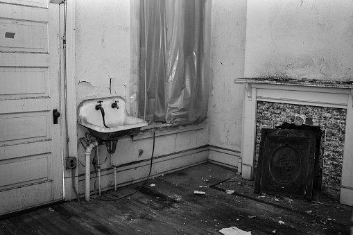 An old residential building had a different use, evidenced by an old ceramic sink installed next to a fireplace.  A window is covered in plastic and trash is on the floor.