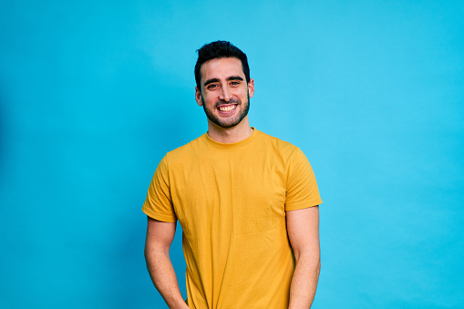 Positive young bearded male in yellow shirt smiling and looking at camera while standing against blue background