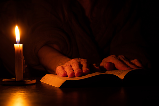 grandmother's hands lie on an open book on the table by the light of a candle in the dark, reading a prayer with a candle