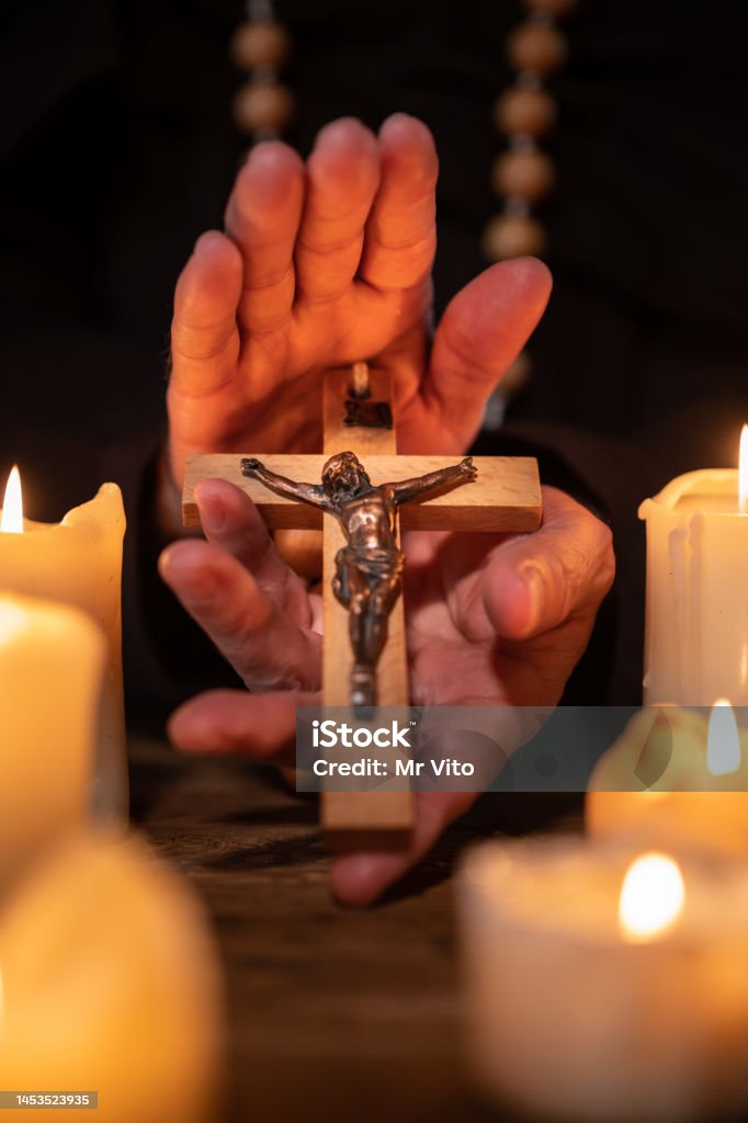 A priest with a cross in his hand In the candlelight, the priest n a brown priest's robe holds in his hands a wooden rosary with a wooden cross and Christ on it. 55-59 Years Stock Photo