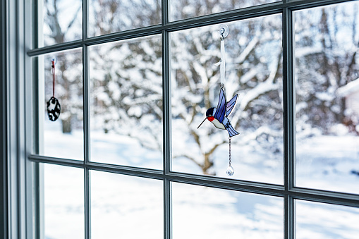 View outside to the snowy winter back yard beyond two hummingbird knick-knack trinkets hanging on the dining room back wall multi-pane bay window glass.