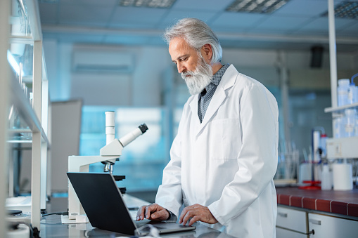 Portrait of a confident Caucasian senior researcher writing down the results. A male scientist using his computer during working in a lab.