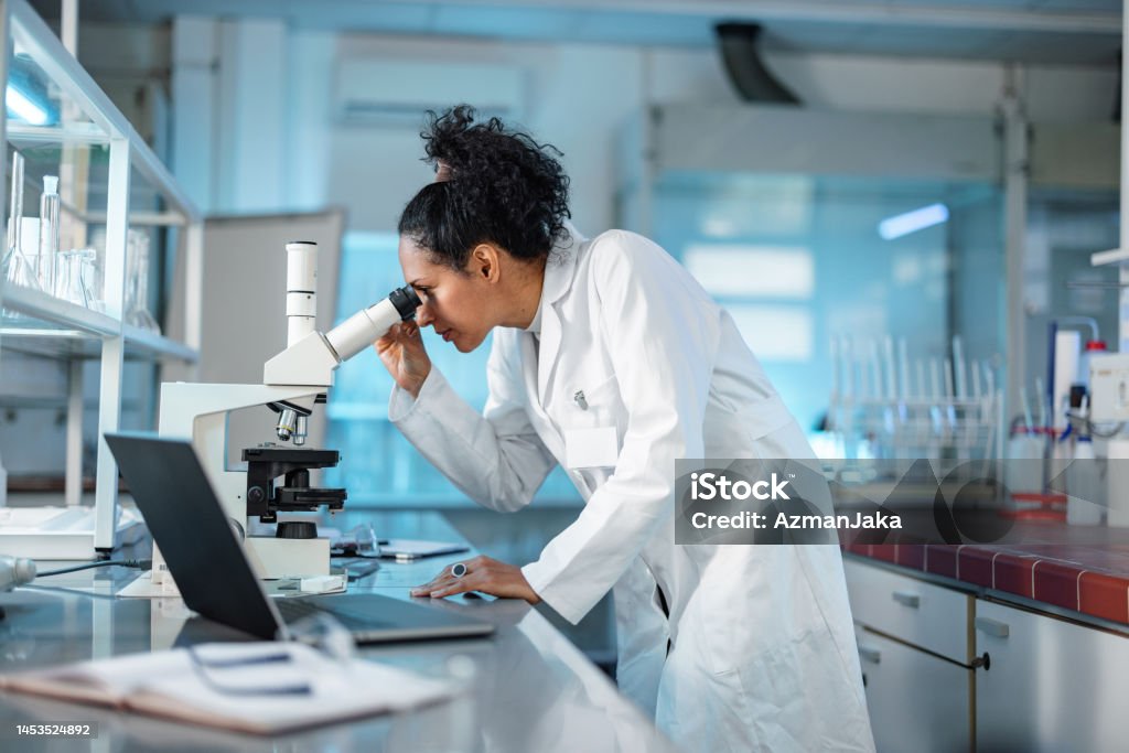 Female Scientist Looking Under Microscope And Using Laptop In A Laboratory Young Hispanic scientist wearing a lab coat, looking under microscope while using laptop in a laboratory. Science Stock Photo