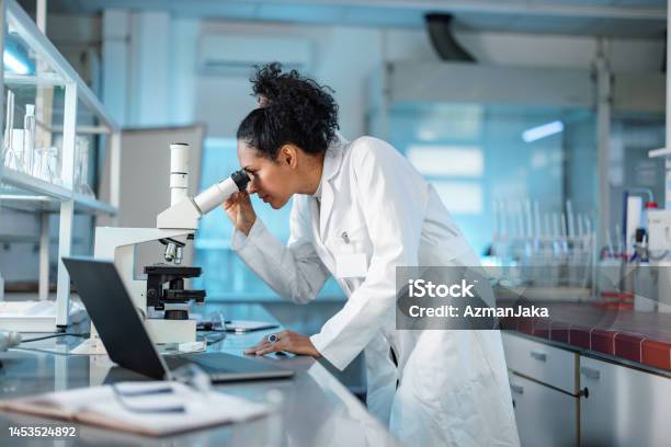 Female Scientist Looking Under Microscope And Using Laptop In A Laboratory Stock Photo - Download Image Now
