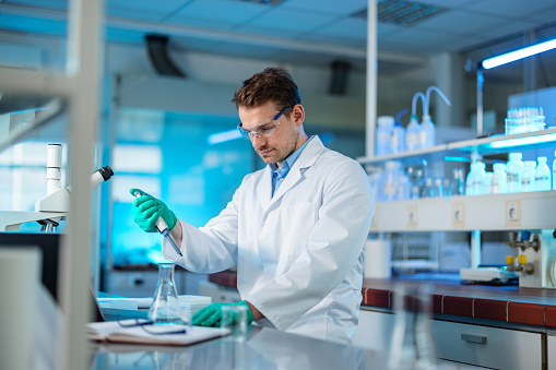 A young adult Caucasian biochemist in his 30s wearing a lab coat and protective glasses. He is mixing liquid chemicals using a pipette in a modern lab.