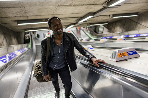 Full length shot of a cheerful mature African American man standing on the escalator in the subway metro station. He is wearing casual clothes and smiling.