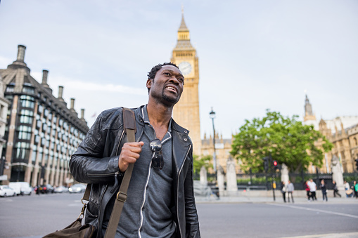Waist up low angle view of a good looking African American man with cool attitude walking in front of the Big Ben in London. He is smiling and looking away.