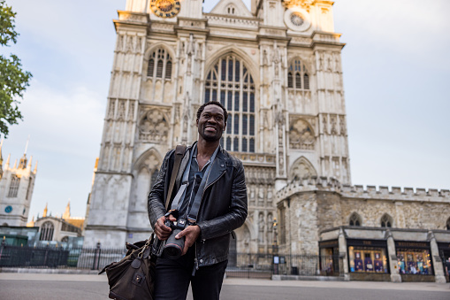 Smiling African American male traveling and photographing in London. He is wearing a black leather jacket and holding a professional camera.