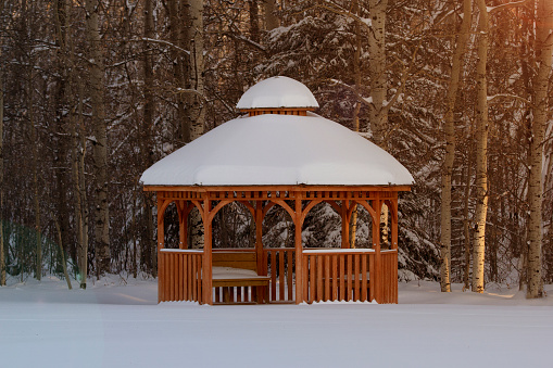 Rustic scene of the wooden cedar brown gazebo in the winter forest covered with snow, warm sunset light.