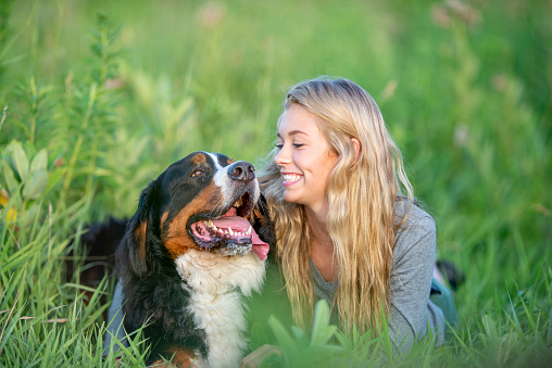 A young blond haired girl lays in the grass with her dog as she poses for a portrait on a warm sunny day.  She is dressed casually and is smiling as the two enjoy the sunshine and warm weather together.