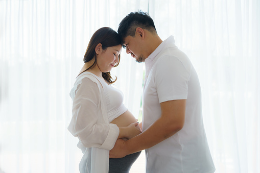 Happy Asian couple expecting baby standing together against window at home, loving husband tenderly touching belly of his pregnant wife