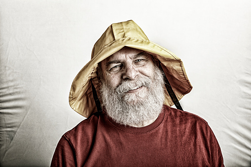 Cheerful salty old man of the sea portrait. Could be a lighthouse keeper? Might be a fisherman fishing boat captain? Maybe just an old gray beard guy with a retro sailor's yellow monsoon rain hat? Deliberate darkened grainy and grubby post treatment.