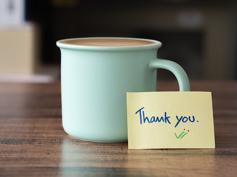 Handwritten text thank you and hot latte coffee in light green ceramic mug on brown wooden table.