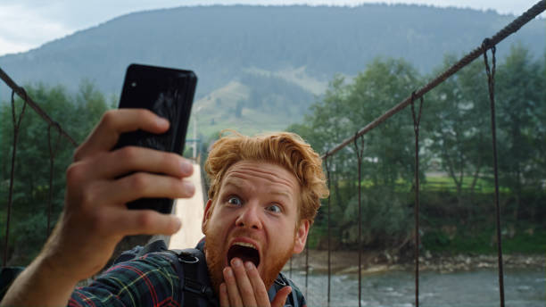 Scared blogger taking selfie on river bridge. Closeup traveler grimace on phone. Scared blogger taking selfie on river bridge. Closeup red hair traveler grimace on mobile phone camera. Emotional guy posing hold cellphone digital device at mountains. Having fun holiday concept. slow motion face stock pictures, royalty-free photos & images